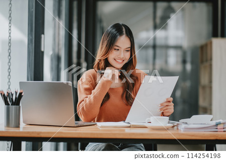 Smiling young Asian businesswoman analyzing documents with a laptop open, in a well-lit modern workspace. Smiling young Asian businesswoman analyzing documents with a laptop open, in a well-lit modern workspace. 114254198