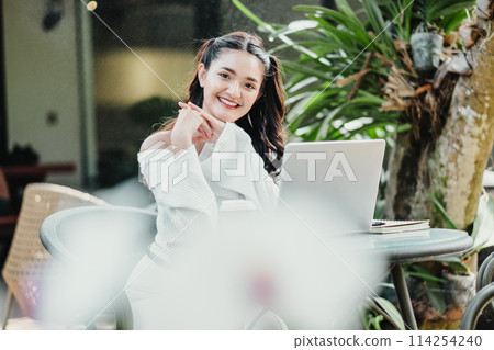 Happy young woman enjoying work on her laptop at a cozy outdoor cafe surrounded by plants. Happy young woman enjoying work on her laptop at a cozy outdoor cafe surrounded by plants. 114254240