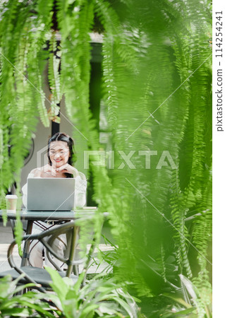 Cheerful woman working on her laptop, framed by vibrant hanging ferns at a garden cafe. Cheerful woman working on her laptop, framed by vibrant hanging ferns at a garden cafe. 114254241