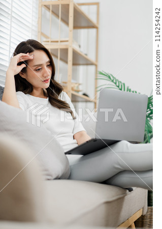 Thoughtful woman seated on a couch, working on a laptop in a well-lit, cozy home setting. Thoughtful woman seated on a couch, working on a laptop in a well-lit, cozy home setting. 114254242