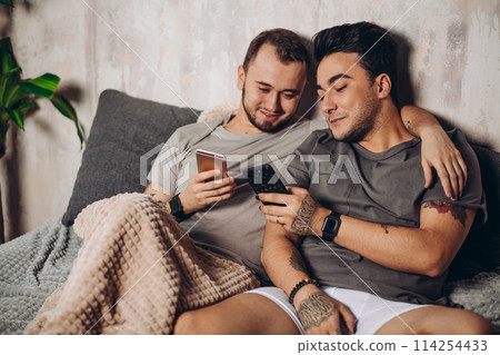 Happy gay couple lying together in bed and using smartphone, looking at photos 114254433