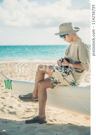 A young man plays the ukulele on a sandy Hawaiian beach with the turquoise ocean in the background A young man plays the ukulele on a sandy Hawaiian beach with the turquoise ocean in the background 114256795