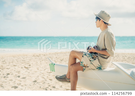 A young man plays the ukulele on a sandy Hawaiian beach with the turquoise ocean in the background 114256799