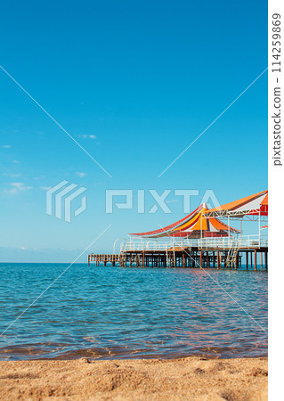 Wooden pier with orange-red awning over water of sea, lake, ocean with beach without people. Travel, vacation, summer background. Vertical. Copy space 114259869