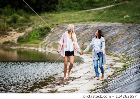 Adult daughter spending time with her mother. Mom and daughter outdoors, walking barefoot in water of lake reservoir, lake embankment. Unconditional, deep maternal love, Mother's Day. 114262657