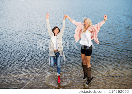Adult daughter spending time with her mother, having fun, jumping. Mom and daughter outdoors, on walk by reservoir, lake embankment. Unconditional, deep maternal love, Mother's Day concept.. 114262663