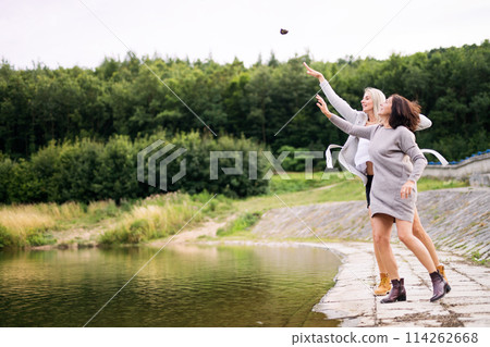 Mom and daughter outdoors, on walk by reservoir, lake embankment, throwing rocks into water. Unconditional, deep maternal love, Mother's Day. 114262668