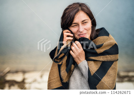 Portrait of beautiful mature woman with blanket around shoulders, standing by lake. Portrait of beautiful mature woman with blanket around shoulders, standing by lake. 114262671
