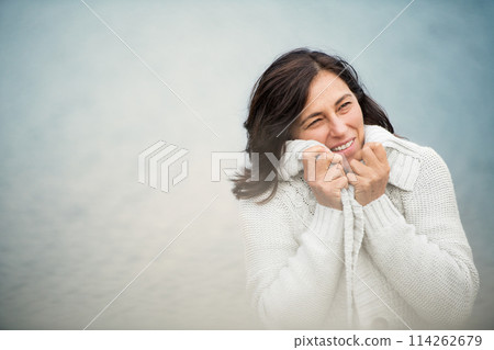 Portrait of beautiful mature woman holding sweater cardigan tightly around her body, standing by lake. Portrait of beautiful mature woman holding sweater cardigan tightly around her body, standing by lake. 114262679