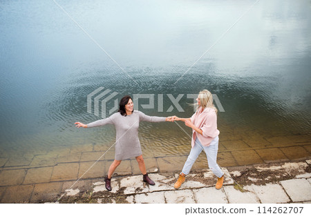 Adult daughter spending time with her mother, having fun, laughing. Mom and daughter outdoors, on walk by reservoir, lake embankment. Unconditional, deep maternal love, Mother's Day concept. Adult daughter spending time with her mother, having fun, laughing. Mom and daughter outdoors, on walk by reservoir, lake embankment. Unconditional, deep maternal love, Mother's Day concept. 114262707