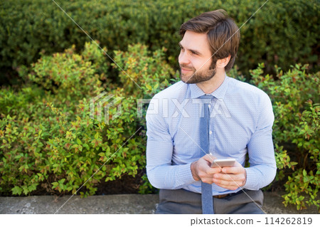 Handsome businessman holding smartphone sitting on bench in city park. Working remotely, waiting for business meeting. Manager smiling, outdoor in urban setting 114262819