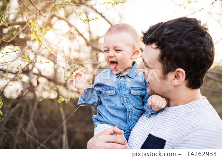 Portrait of father holding little toddler boy, showing him buds on twig. Family time oudroor during warm spring day. Father's day concept. 114262933