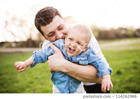 Father holding little toddler boy kissing him, making him laugh, having fun during warm spring day. Father's day concept. 114262966