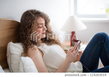 Beautiful woman with curly hair sitting on bed, scolling on smartphone. 114263014
