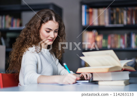 Young female student in library, focusing on final project, presentation. University student preparing for final exam. 114263133