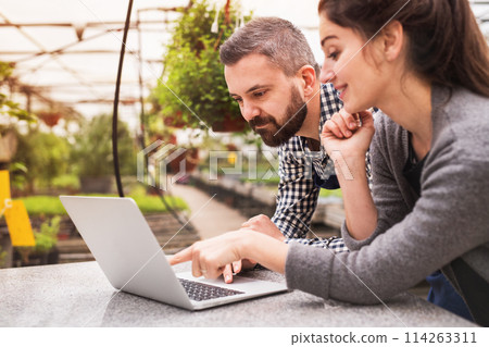 Female business owner and gardener looking at order in laptop, preparing flowers and seedlings for customer. Small greenhouse business. Female business owner and gardener looking at order in laptop, preparing flowers and seedlings for customer. Small greenhouse business. 114263311