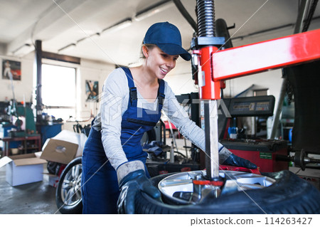 Female auto mechanic changing tieres in auto service, using machinery to removing tire. Beautiful woman working in garage. 114263427