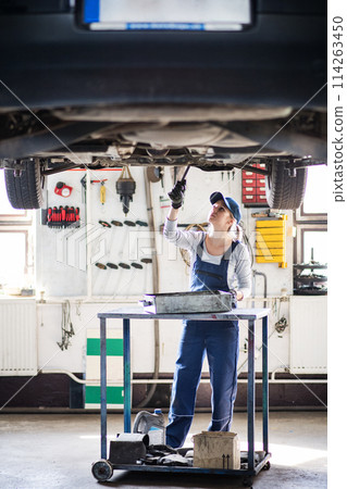 Female auto mechanic elevating car on car lift, changing oil. Beautiful woman working in a garage, wearing blue coveralls. 114263450