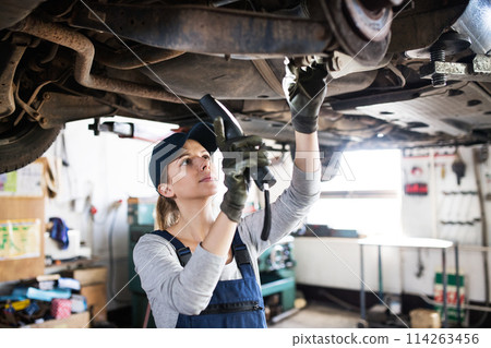 Female auto mechanic elevating car on car lift, working underneath. Beautiful woman working in a garage, wearing blue coveralls. 114263456