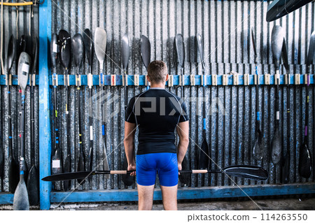 Rear view of young canoeist standing in front of wall with paddles. Concept of canoeing as dynamic and adventurous sport. Rear view of young canoeist standing in front of wall with paddles. Concept of canoeing as dynamic and adventurous sport. 114263550