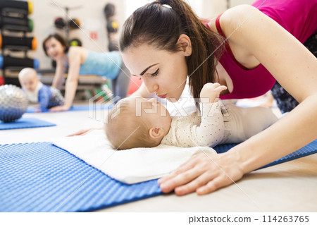 Mother smiling at baby while performing push-ups, kissing infant. Group exercise class, fitness or pilates for parents. Moms staying active while boding with babies. Mother smiling at baby while performing push-ups, kissing infant. Group exercise class, fitness or pilates for parents. Moms staying active while boding with babies. 114263765