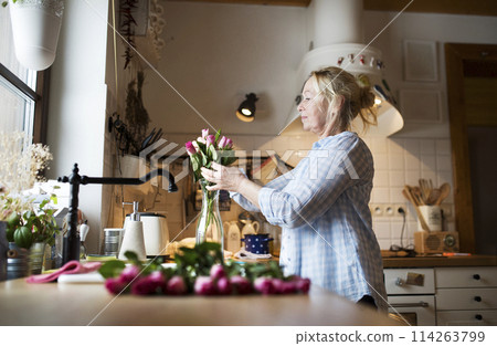Beautiful mature woman arranging bouquet of roses in a vase, a hobby and relaxation. Older woman living alone, enjoying peaceful weekend day Beautiful mature woman arranging bouquet of roses in a vase, a hobby and relaxation. Older woman living alone, enjoying peaceful weekend day 114263799