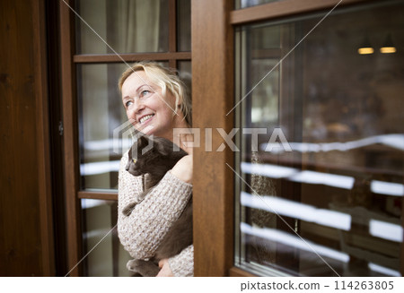 Beautiful mature woman at home, holding, petting her cat, looking from window. Older woman living alone, enjoying peaceful weekend day Beautiful mature woman at home, holding, petting her cat, looking from window. Older woman living alone, enjoying peaceful weekend day 114263805