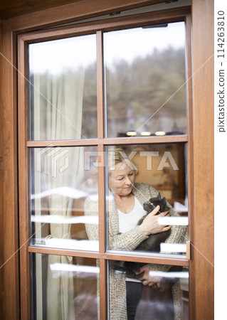 Beautiful mature woman at home, holding, petting her cat, looking through the window. Older woman living alone, enjoying peaceful weekend day Beautiful mature woman at home, holding, petting her cat, looking through the window. Older woman living alone, enjoying peaceful weekend day 114263810