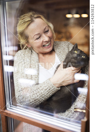 Beautiful mature woman at home, holding, petting her cat, looking through the window. Older woman living alone, enjoying peaceful weekend day Beautiful mature woman at home, holding, petting her cat, looking through the window. Older woman living alone, enjoying peaceful weekend day 114263812