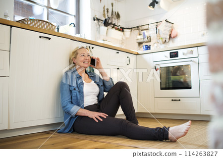 Beautiful mature woman at home, phone calling, sitting on floor in kitchen. Older woman living alone, enjoying peaceful weekend day Beautiful mature woman at home, phone calling, sitting on floor in kitchen. Older woman living alone, enjoying peaceful weekend day 114263827
