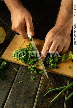 Slicing fresh parsley with a knife in the hands of a chef on a cutting board. Low key concept of preparing vegetable salad. Advertising space 114264495