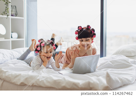 Woman lying with a cute girl on the bed, curling their hair, using a laptop. Woman lying with a cute girl on the bed, curling their hair, using a laptop. 114267462