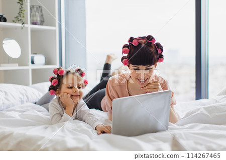 Woman lying with a cute girl on the bed, curling their hair, using a laptop. Woman lying with a cute girl on the bed, curling their hair, using a laptop. 114267465