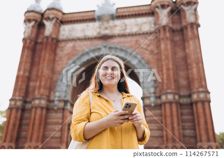 Woman with smart phone in summer vacation travel, standing near Barcelona triumphal arch. Female traveler visiting local landmarks. Concept of travel, tourism and vacation in city Woman with smart phone in summer vacation travel, standing near Barcelona triumphal arch. Female traveler visiting local landmarks. Concept of travel, tourism and vacation in city 114267571