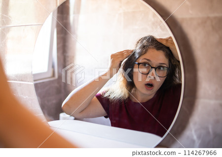 Gray haired surprised caucasian middle aged woman looking at grey hair head in mirror reflection. Early gray hair concept 114267966