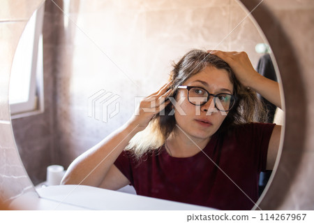 Gray haired surprised caucasian middle aged woman looking at grey hair head in mirror reflection. Early gray hair concept Gray haired surprised caucasian middle aged woman looking at grey hair head in mirror reflection. Early gray hair concept 114267967