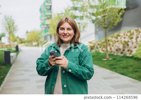 30s cheerful woman tourist dressed in casual look holding smartphone gadget in hand for communicate on the city street. Happy woman chatting on phone and relaxing outdoors. Urban, people concept. 114268596