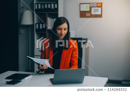 Smiling young Asian businesswoman working on a laptop computer at her desk in a bright modern home office, doing calculating expense financial report finance making notes on paper graph data document. Smiling young Asian businesswoman working on a laptop computer at her desk in a bright modern home office, doing calculating expense financial report finance making notes on paper graph data document. 114269352