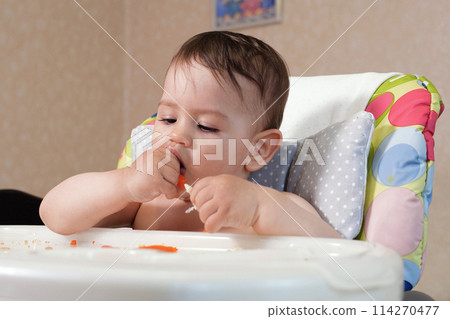 Portrait of little baby boy eating food. Baby with a spoon in feeding chair. Cute baby eating first meal 114270477