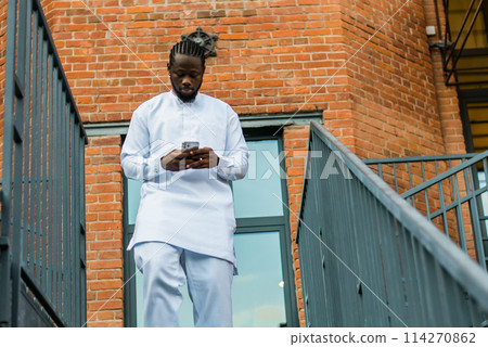 African american man in stylish ethnic dashiki wear viewing photos in social networks on mobile enjoying free time outdoors. Dark-skinned man watching funny video on smartphone and social networks 114270862