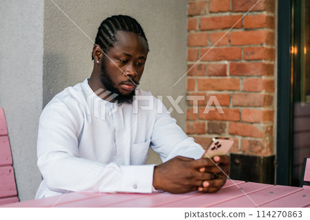 African american man in stylish ethnic dashiki wear viewing photos in social networks on mobile enjoying free time outdoors. Dark-skinned man watching funny video on smartphone and social networks 114270863