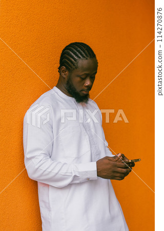 African american man in stylish ethnic dashiki wear viewing photos in social networks on mobile enjoying free time outdoors. Dark-skinned man watching funny video on smartphone and social networks 114270876
