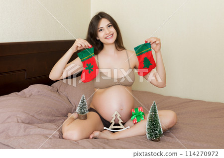 Cute happy pregnant woman in pajamas at home with christmas decoration. Mother to be with big belly, hands over tummy. Merry Christmas holidays. New Year Eve concept 114270972