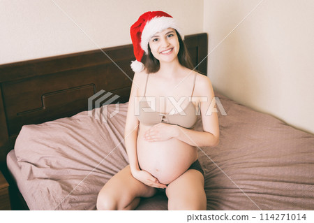 Cute happy pregnant woman in pajamas at home in santa hat. Mother to be with big belly, hands over tummy. Merry Christmas holidays. New Year Eve concept Cute happy pregnant woman in pajamas at home in santa hat. Mother to be with big belly, hands over tummy. Merry Christmas holidays. New Year Eve concept 114271014