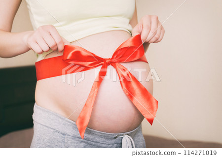 Belly of a pregnant woman tied with a red bow on a home background Belly of a pregnant woman tied with a red bow on a home background 114271019