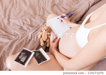 Top view caring future mother writing diary with ultrasound. pregnant woman tummy making notes feeling during pregnancy or creating scrapbook 114271070