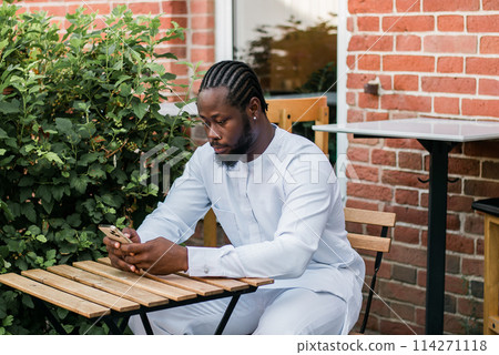 African american man in stylish ethnic dashiki wear viewing photos in social networks on mobile enjoying free time outdoors. Dark-skinned man watching funny video on smartphone and social networks African american man in stylish ethnic dashiki wear viewing photos in social networks on mobile enjoying free time outdoors. Dark-skinned man watching funny video on smartphone and social networks 114271118