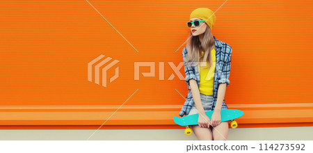 Summer portrait of stylish young woman with skateboard in colorful clothes on orange background 114273592