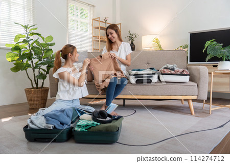 Young Asian woman packing clothes to the suitcase. Preparation for the summertime vacation. Two women are planning a trip and helping to prepare luggage to travel Young Asian woman packing clothes to the suitcase. Preparation for the summertime vacation. Two women are planning a trip and helping to prepare luggage to travel 114274712