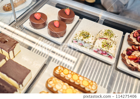 A variety of cakes covered with chocolate and decorated with pistachio mousse. A showcase in a pastry shop with delicious desserts with fresh berries and cream. Delicious pastries in the coffee shop.  114275406
